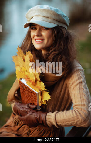 Bonjour l'automne. Portrait of an old woman 40 moderne en pull, jupe, chapeau, gants et écharpe avec ordinateur portable et feuilles jaunes tout en restant assis sur un b Banque D'Images