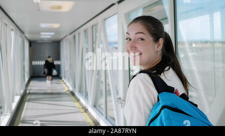 Happy young woman with backpack est passe passerelle télescopique à l'avion. Elle se retourne, regarde la caméra et sourit. Banque D'Images