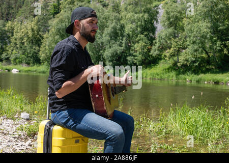 Jeune homme barbu hippie, dans un bouchon joue de la guitare dans le contexte d'un paysage d'été, ciel bleu et les montagnes. le transport des prises Banque D'Images