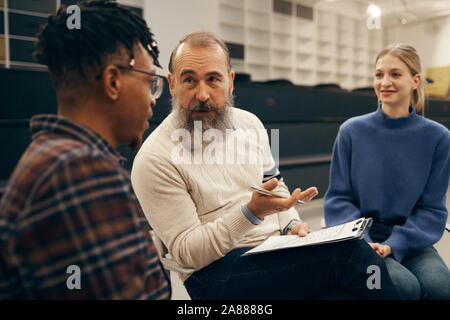 Man avec barbe, assis sur une chaise avec le presse-papiers et de parler aux jeunes pendant la leçon Banque D'Images