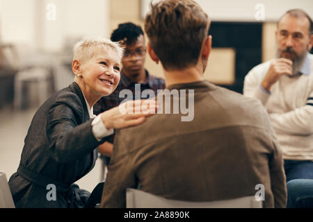 Smiling mature woman avec de courts cheveux blonds de parler au jeune homme qui, assis sur la chaise retour à l'appareil photo pendant la classe thérapeutique Banque D'Images