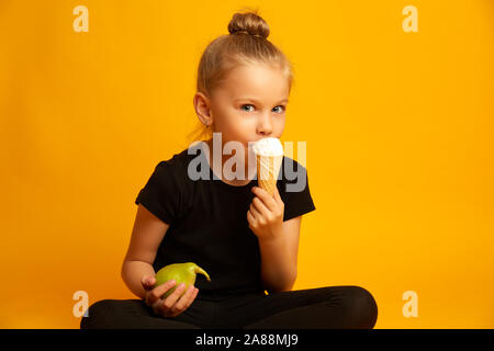 Ahuri petite fille en collants et chaussures de danse choisir entre des aliments sains et malsains, en position assise, les jambes croisées sur fond jaune Banque D'Images