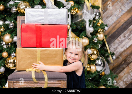 Portrait de jeune fille d'âge élémentaire portant robe bleue à côté de la tenue de l'arbre de Noël gros tas de cadeaux emballés en toute sécurité avec une assurance d'obtenir beaucoup de présent Banque D'Images