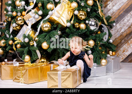 Portrait de jeune fille d'âge élémentaire portant robe bleue assise à côté de grand déballage de l'arbre de Noël cadeau enveloppé fort curieux à propos d'un présent le lendemain de Banque D'Images