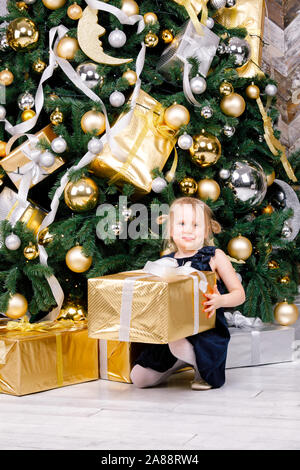 Portrait de jeune fille d'âge élémentaire portant robe bleue assise à côté arbre de Noël cadeau enveloppé holding big fort heureux d'obtenir un présent le lendemain de Banque D'Images