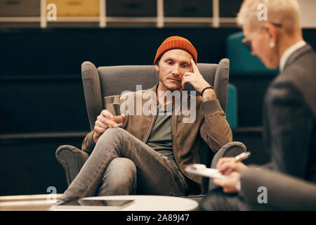 Jeune homme sérieux en vêtements décontractés sitting on sofa l'eau potable et à l'attente de la conclusion de la médecin Banque D'Images