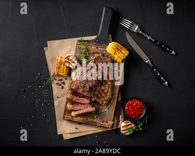 Tranchés, Fried steaks juteux avec des herbes et épices dispersés sur une planche en bois, parchemin, l'ail et le maïs grillé, sauce dans un petit bol, fo acier foncé Banque D'Images