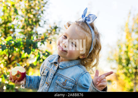 Cute little girl enfant manger bio mûre pomme rouge dans le verger en automne. Juste curly haired fille européenne enfant dans un costume en jean à la ferme Banque D'Images