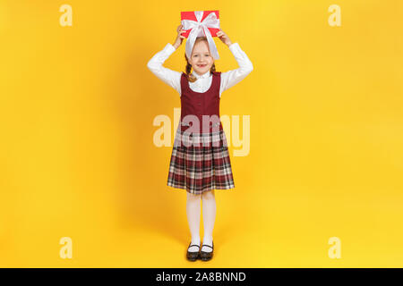 Funny little girl holding une boîte avec un cadeau sur sa tête. Un enfant dans un long avec tresses dans une chemise blanche et une robe sur un cri de Bourgogne Banque D'Images