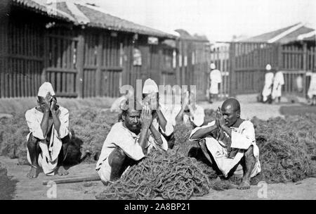 La photographie montre cinq hommes prisonniers accroupis avec mains pressées ensemble au milieu d'un matériau fibreux à Rangoon, Birmanie 1910-1920 Banque D'Images