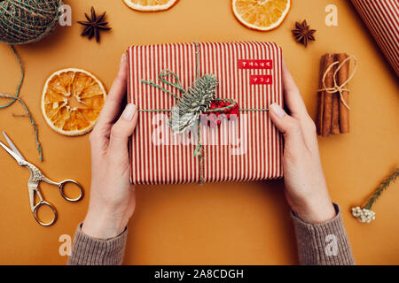 Close-up of woman holding Christmas present qu'elle fait par elle-même pour Noël et décoré avec des branches de sapin et de baies sur fond jaune Banque D'Images