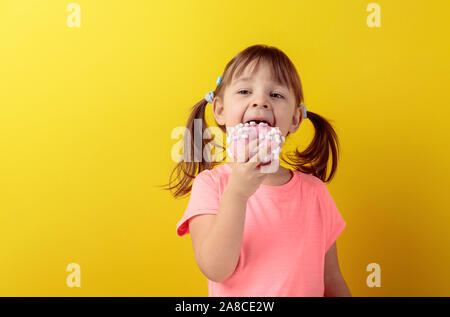 Fillette de quatre ans dans un t-shirt rose eat donut. Les cheveux de la fille est liée à la queue. Fond jaune. Banque D'Images