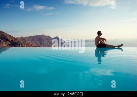 Jeune homme solitaire en appui sur le bord d'une piscine à débordement surplombant la Méditerranée pittoresque vue sur la caldeira de Santorin, Grèce Banque D'Images