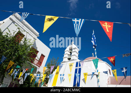 Vue ensoleillée et lumineuse sur la plaza décorée avec un drapeau pennant représentant la Grèce, l'Empire byzantin et l'église orthodoxe grecque de Mykonos Banque D'Images