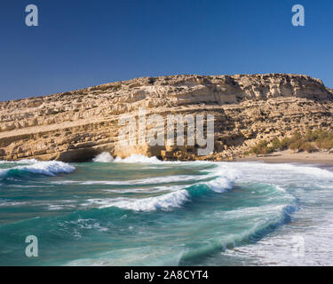 Matala, Héraklion, Crète, Grèce. Le martèlement des vagues plage en contrebas de falaises de grès, l'ancienne maison d'habitation troglodyte hippies. Banque D'Images