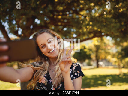 Portrait of a smiling young blonde woman sitting on a mobile phone selfies montrant signe de paix dans le parc par une journée ensoleillée Banque D'Images