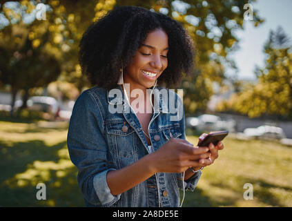 Portrait of a smiling afro-américain à la mode jeune femme textos message sur téléphone mobile debout dans le parc mark d'écoute Banque D'Images