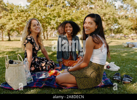 Smiling female friends multiraciale siégeant ensemble sur couverture sur l'herbe verte avec des fruits dans le parc looking at camera Banque D'Images