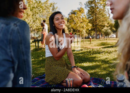 Portrait of a smiling young woman eating multiraciale avec tomate cerise à pique-nique dans le parc lors d'une journée ensoleillée en été Banque D'Images