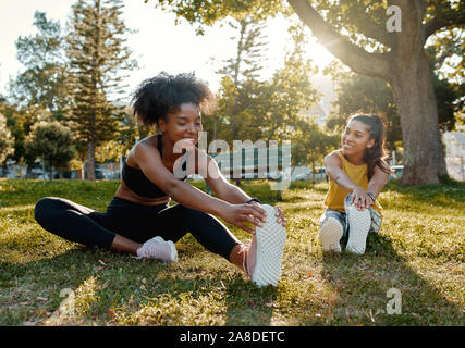 Les jeunes femmes divers amis assis sur l'herbe verte qui s'étend ses jambes dans la lumière du soleil du matin au parc - divers les amis de préchauffage avant de faire Banque D'Images
