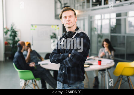 Young Caucasian indépendant avec sourire sur son visage et d'écouteurs en permanent lieu de coworking bureau loft alors que ses collègues assis à la table et Banque D'Images