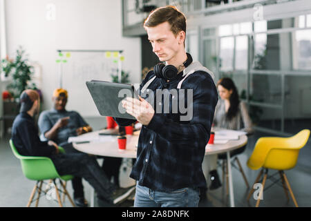 Portrait de demi-longueur cheerful young manager ou le concepteur habillés en tenue décontractée et écouteurs, ayant travailler dans office, holding digital tablet Banque D'Images