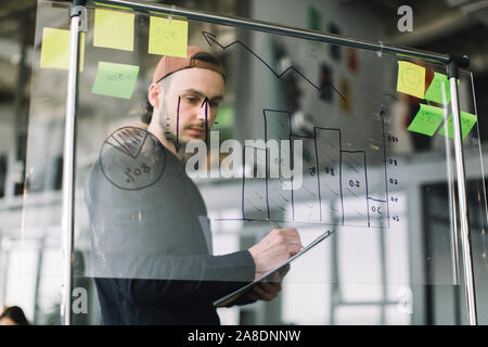 Jeune homme en tenue décontractée et les travailler avec les données et les schémas sur le bureau en verre mur avec les notes, tandis que ses collègues de discuter des idées et Banque D'Images