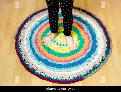 Jeune enfant de 3 ans debout sur un tapis rond en crochet coloré de t-shirts réaffectés sur un plancher de bois brun. Vue de dessus. Banque D'Images