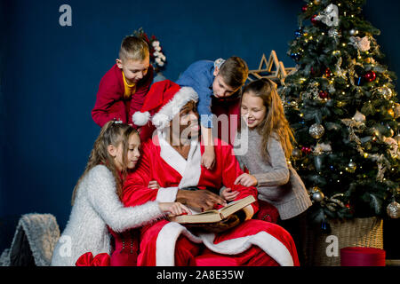 La veille de Noël, le Père Noël lit l'histoire de Noël pour les enfants qui l'écoutent de tous les côtés, sur l'arrière-plan d'un arbre de Noël. Banque D'Images