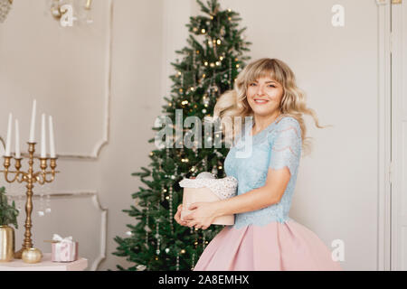 Jeune femme décore le sapin de Noël avec des jouets de Noël. Aménagement intérieur classique en blanc et or Banque D'Images