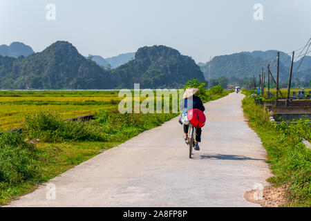 Un résident local Tam Coc rides le long d'une petite route à côté de champs de riz et de magnifiques paysages de montagne à Ninh Binh, Vietnam du Nord - Automne 2019 Banque D'Images