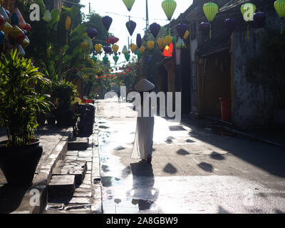 Vietnamienne portant un ao dai ou tunique traditionnelle et pant costume et un chapeau de paille conique de la ou en marchant sous les lanternes dans un tissu de Hoi An Banque D'Images