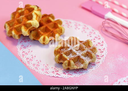 Petites gaufres rondes frais avec du sucre en poudre sur le papier-dentelle napperon rose et sol avec des ustensiles de cuisson en arrière-plan Banque D'Images