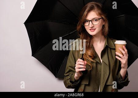 Une fille se tient dans le contexte d'un parapluie noir ouvert dans un trench-coat vert et verres avec une tasse de papier dans sa main. Phot horizontale Banque D'Images