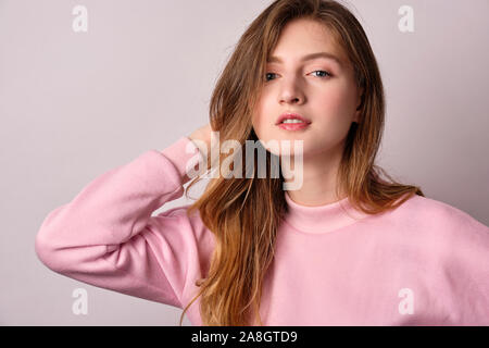 Une fille blonde dans un sweat rose se dresse sur un fond blanc et met sa main derrière sa tête en regardant la caméra, photo horizontale Banque D'Images