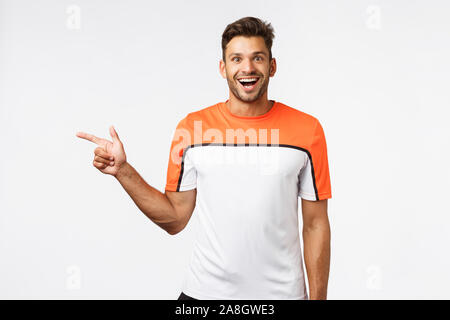 Heureux beau sportif barbu dans sports t-shirt pointant vers la gauche, voir refroidir l'équipement dans une salle de sport, s'est demandé et surpris, étonné de l'appareil photo en souriant Banque D'Images