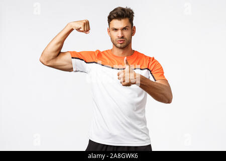 Sassy beau barbu masculin dans le sport t-shirt, tendue, les muscles biceps show et le pouce, faire le fort sérieux, heureux avec une bonne Banque D'Images