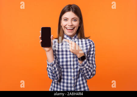 Portrait of attractive brunette woman with charmant sourire portant chemise à carreaux l'article pointant sur téléphone portable, satisfait de services et applications mobiles. Banque D'Images