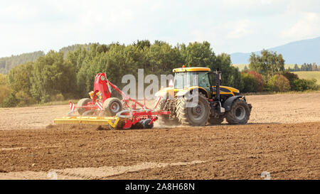 Tracteur jaune rouge tire sur le mécanisme des champ, arbres en arrière-plan. Banque D'Images