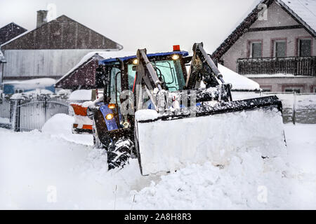 Petit tracteur avec charrue supprimant les charges de neige lors d'une forte tempête de calamité, maisons de village en arrière-plan. Banque D'Images