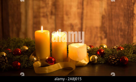 La chaleur de la notion de Noël : trois chandelles allumées sur une table en bois foncé et un cadre rustique avec des branches de pins, des cônes de pin et de l'or et Banque D'Images