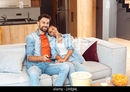 Mixed Race Mariage. Young man and woman hugging regardant la tv programme ainsi des changements de chaînes avec télécommande heureux Banque D'Images