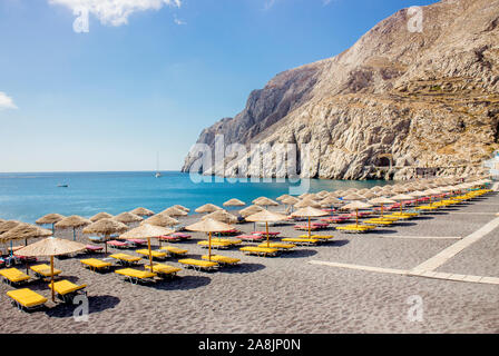 Belle plage de sable noir sur l'île grecque de Santorin, Kamari village, matin ensoleillé. Beaucoup de soleil, chaises longues et parasols en paille une ligne. T Banque D'Images