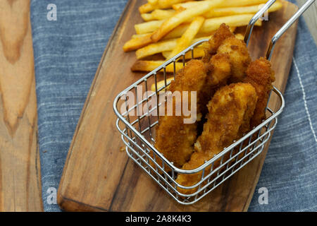 Coujons de poulet enrobées de chapelure dans un fil panier métal, avec des frites, sur une planche à découper en bois Banque D'Images