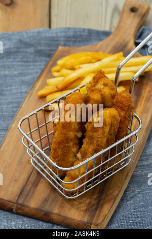 Coujons de poulet enrobées de chapelure dans un fil panier métal, avec des frites, sur une planche à découper en bois Banque D'Images