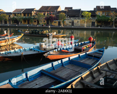 Bateaux de pêche en bois, certaines avec un tissu lanternes, amarré dans le calme de la rivière Thu Bon. Bâtiments en stuc d'or en face de la ligne de rive. Banque D'Images