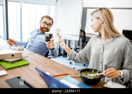 L'homme et de la femme de manger une salade et boire du café pendant une heure du déjeuner sur le lieu de travail sans quitter le bureau Banque D'Images