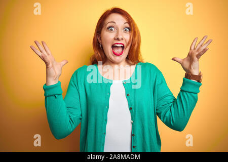 Belle jeune femme rousse portant chandail vert d'hiver isolé sur fond jaune célébrant la folie et surpris du succès avec un bras levés Banque D'Images