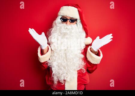 L'âge moyen homme portant un costume de Père Noël et des lunettes de soleil sur fond rouge isolé clueless et expression confus avec les bras et les mains posées. N Banque D'Images