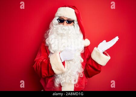 L'âge moyen homme portant un costume de Père Noël et des lunettes de soleil sur fond rouge isolé surpris et souriant à la caméra tout en se présentant avec la main et Banque D'Images